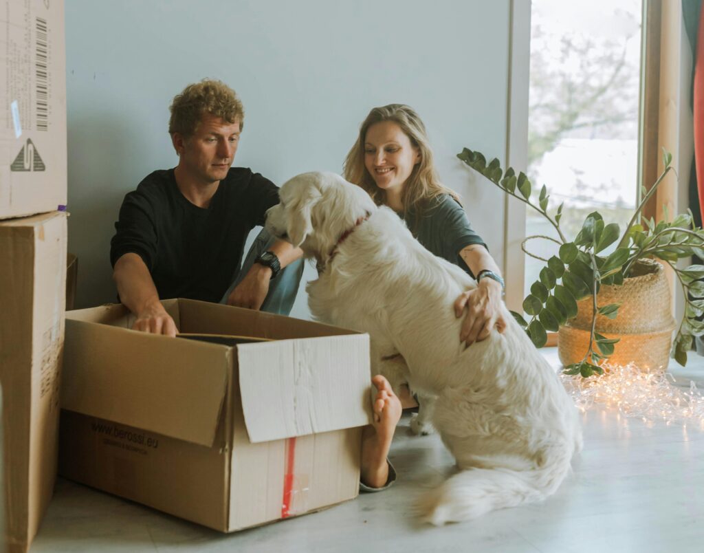 A man and a woman petting their dog surrounded by moving boxes in a new home.
